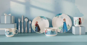 Vaisselle et ustensiles pour enfants à l'effigie des personnages de La Reine des neiges, exposés sur une étagère bleue. Vaisselle et ustensiles pour enfants à l'effigie des personnages de La Reine des neiges, présentés dans des coffrets cadeaux bleus sur une étagère bleue.