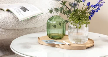 Une table basse ronde avec un vase Villeroy & Boch Fleur contenant des fleurs bleues et vertes. Une table basse ronde avec un vase Villeroy & Boch Fleur contenant des fleurs bleues et vertes. En arrière-plan, un magazine ouvert est posé sur une chaise blanche et moelleuse.