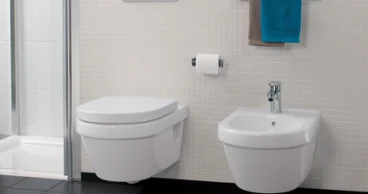 Une salle de bains moderne Architectura avec des WC suspendus, un bidet, un porte-papier toilette et un porte-serviettes avec une serviette bleu et marron.  Le sol est recouvert de carrelage noir et le mur de petits carreaux blancs carrés.