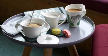 Une tasse à café NewWave, un pot à lait, un sucrier avec une cuillère et deux macarons sur une sous-tasse, sur une petite table.