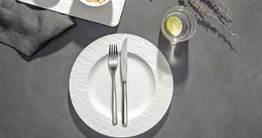 Couverts Piemont sur une assiette Manufacture Une assiette avec une fourchette croisée et un couteau dessus, à côté d'un verre d'eau avec du citron vert et deux bols de snacks sur une table.