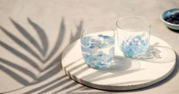 Deux verres à eau Like Aqua sur un plateau rond Deux verres à eau Like Aqua avec des pétales bleus sur un plateau rond, avec des ombres de feuilles de palmier sur une surface claire.