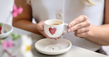 Une personne tient une tasse à café blanche avec un motif cœur rouge au-dessus d'une soucoupe assortie avec motif cœur rouge posée sur une table.