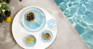 Un plateau rond avec de la vaisselle de la collection Perlemor Aqua et un verre d'eau, au bord d'une piscine.
