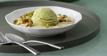 Une coupe à dessert Pura avec de la glace à la pistache Une coupe à dessert Pura contenant de la glace à la pistache, des pistaches hachées et de la crème, posée sur une assiette blanche avec des couverts à côté.