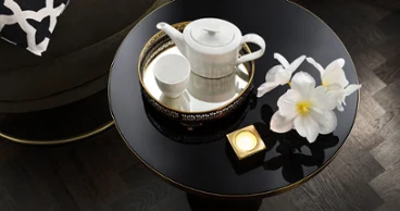 Une théière MetroChic blanche avec une tasse assortie, deux fleurs blanches et une bougie allumée sont posées sur une table ronde noire avec un coussin à motifs en arrière-plan.