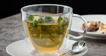 Une tasse Artesano Hot&Cold Beverages avec des feuilles de menthe sur une sous-tasse, accompagnée d’une cuillère, sur un fond sombre.