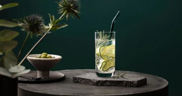 Un verre La Divina de Villeroy & Boch servi avec style Un verre La Divina avec des tranches de concombre et du romarin, sur une surface en pierre avec un fond vert. Une paille en verre Artesano est plantée dans la boisson et une coupe d’olives est posée à côté.