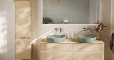 Une salle de bains bohème avec des WC et un bidet Une salle de bains bohème moderne de Villeroy & Boch avec des WC muraux Venticello et un bidet sur un sol en galets. Une plante décorative et un panier en osier sont posés sur un rebord en bois au-dessus.