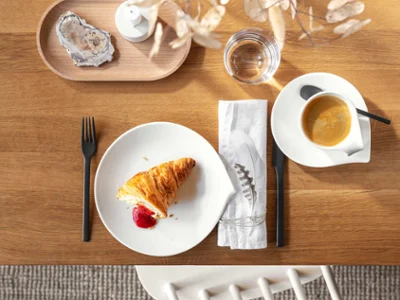 Une table dressée pour le petit-déjeuner avec vaisselle Flow, vue du dessus Vue de dessus d’une table en bois avec un croissant et de la confiture sur une assiette Flow blanche, une tasse à café, des couverts de table, une serviette, un verre d’eau et des éléments décoratifs.