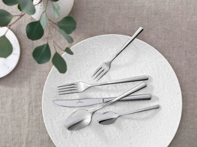 Des couverts de table Piemont avec une assiette Manufacture blanche sur une nappe neutre Une décoration de table élégante avec des couverts de table Piemont modernes et de verdure décorative sur une nappe neutre.