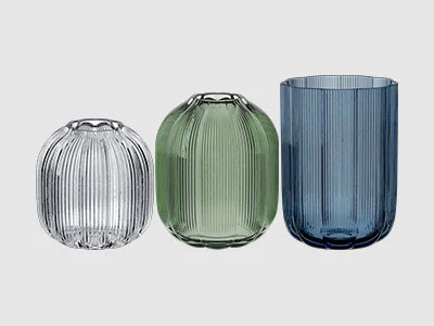 Image de la collection - Vases Fleur Trois vases en verre strié de différentes hauteurs et couleurs : transparent, vert clair et bleu foncé, disposés côte à côte.