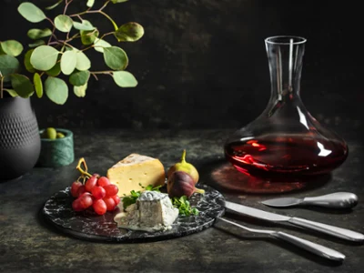 Un décanteur à vin rouge Maxima sur une surface structurée foncée Un plateau de fromage gourmet avec des raisins et des figues à côté d’un décanteur à vin rouge Maxima sur une surface structurée sombre.