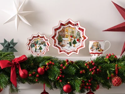 La collection Toy’s Fantasy de Villeroy & Boch Une collection Toy’s Fantasy de Villeroy & Boch, une assiette décorative, un gobelet orné de paysages hivernaux, des ornements en forme d’étoile et une guirlande luxuriante sur le manteau de la cheminée, décorée de rubans rouges et de baies.