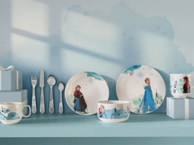 Vaisselle et ustensiles pour enfants à l'effigie des personnages de La Reine des neiges, exposés sur une étagère bleue. Vaisselle et ustensiles pour enfants à l'effigie des personnages de La Reine des neiges, présentés dans des coffrets cadeaux bleus sur une étagère bleue.