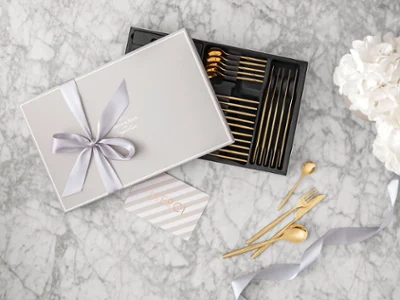 Une boîte cadeau avec des couverts de table MetroChic dorés et un ruban Une boîte cadeau avec des couverts de table MetroChic dorés et un ruban, une carte de remerciement et des fleurs blanches sur une surface en marbre.