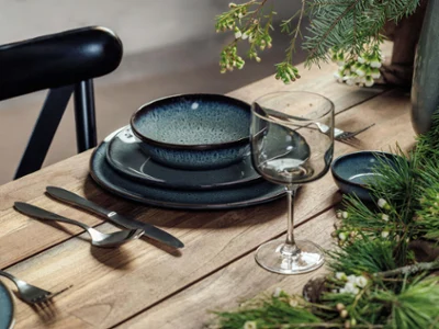De la vaisselle Crafted et un verre Winter Glow sur une table de Noël Une table en bois est dressée avec un verre Winter Glow ainsi que des assiettes et des plats creux en céramique Crafted bleu foncé. Des plantes vertes et des fleurs blanches décorent la table.