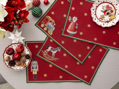 Sets de table Gobelin Toy's Delight aux motifs de Noël Vue de dessus de quatre sets de table rouges en tapisserie Gobelin Toy's Delight ornés d'un casse-noisette, d'un cheval à bascule, d'un train et du Père Noël, disposés sur une table décorée de biscuits de Noël, de pommes et de noix