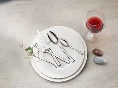 Un ensemble de couverts de table Victor sur une assiette Cellini blanche Un ensemble de couverts Victor avec un couteau, une fourchette et deux cuillères est posé sur des assiettes Cellini blanches, à côté d’un verre à vin rouge et de deux pierres lisses sur une surface en marbre.
