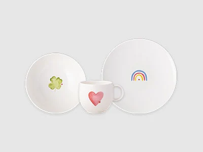 Image de la collection - With Love Trois assiettes blanches : une assiette avec un arc-en-ciel, un bol avec une fleur verte et une tasse avec un cœur rose sur un fond gris.