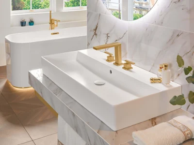 Une robinetterie de lavabo dorée Mettlach avec lavabo et baignoire Une salle de bains moderne avec des surfaces en marbre, un grand lavabo rectangulaire, des robinetteries dorées, un miroir rond, une baignoire en îlot et une plante verte en guise de décoration. La lumière naturelle passe à travers les fenêtres.