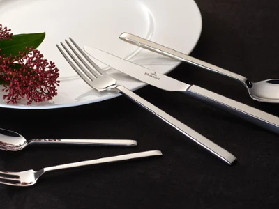 Des couverts de table La Classica sur une assiette blanche avec une fleur décorative Des couverts de table La Classica brillants et une fleur décorative sur une assiette blanche devant un fond sombre.