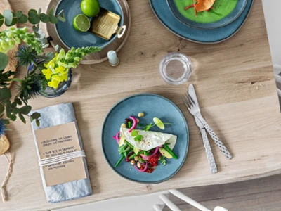 Couverts Boston avec une assiette Lave Une assiette bleue avec de la salade fraîche, des couverts, une serviette et des articles de table décoratifs sur une table en bois.