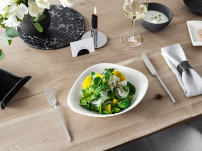 Des assiettes Vapiano avec de la salade et des couverts de table de NewWave Une coupe Vapiano de Villeroy & Boch avec une salade sur une table en bois, à côté d’un couteau et d’une fourchette NewWave.