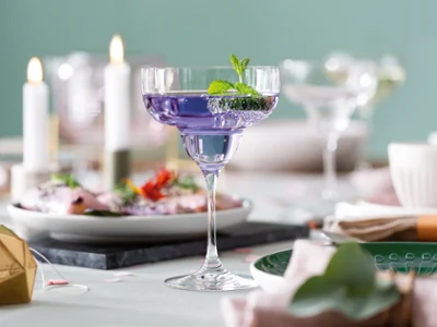 Un verre Purismo avec cocktail violet Un cocktail violet, garni de menthe, servi dans un verre à margarita Purismo sur une table dressée, entourée d’assiettes, de bougies et d’objets décoratifs.