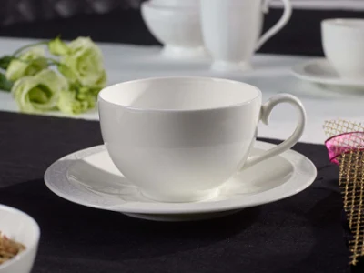 Une tasse White Pearl avec sous-tasse Une tasse et sous-tasse blanches en porcelaine sur une nappe noire, avec des tasses blanches floues en arrière-plan et une fleur décorative.