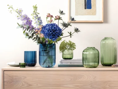 Vases à fleurs sur une commode marron Vases en verre vert et bleu de différentes formes, certains avec des fleurs à l'intérieur, posés sur un meuble en bois avec une photo sur le mur derrière eux