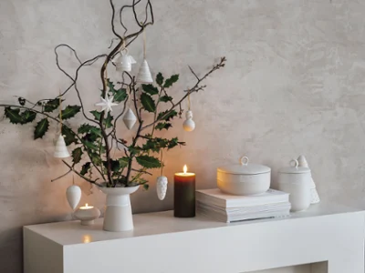 Une décoration de Noël moderne sur une étagère blanche Une décoration de Noël moderne et minimaliste sur un présentoir blanc avec un vase Winter Glow, des branches et des ornements suspendus.