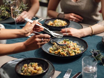 Sur une nappe bleue, des plats creux Lave remplis de salades composées. Quatre personnes profitent d’un repas à une table. Sur une nappe bleue, des plats creux Lave remplis de salades composées.