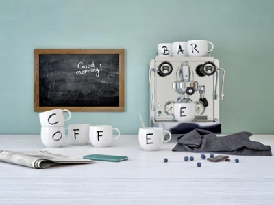 Des mugs de la collection Letters à côté d’une machine à café Une machine à expresso avec des tasses « Letters » de Villeroy & Boch épelant le mot « COFFEE » (CAFÉ), sur une table, à côté d’un téléphone, d’un journal, de myrtilles et d’un tableau portant l’inscription « Guten Morgen ! » (Bonjour).