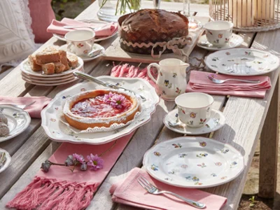 De la vaisselle Petite Fleur sur une table dressée pour le café La collection Petite Fleur de Villeroy & Boch est élégamment disposée sur une table en bois et comprend des assiettes à motifs floraux, un gâteau, une tarte, des tasses et des serviettes roses pour le thé.