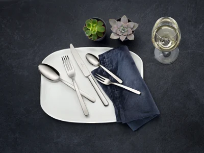 Des couverts de table Louis sur une assiette blanche avec une serviette bleue Une table dressée avec une assiette blanche, des couverts de table en acier inoxydable de la collection Louis, une serviette foncée, deux petites succulentes et un verre à vin blanc sur une surface foncée.