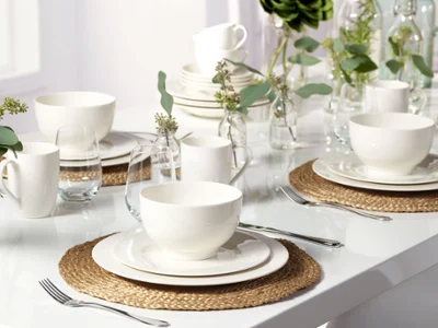 De la vaisselle Basic White de Villeroy & Boch sur des sets de table tissés avec des vases décoratifs et des branches vertes Un élégant ensemble de vaisselle blanche de la collection Basic White de Villeroy & Boch sur une table avec des sets de table décoratifs et des plantes vertes.
