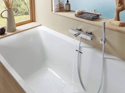 Un thermostat de baignoire Universal Taps & Fittings avec une baignoire blanche et une douchette à main Une salle de bains moderne avec une baignoire blanche, des touches de bois et de grande fenêtre. La baignoire est équipée d’un robinet chromé et d’une douchette à main. Sur le rebord de la fenêtre, il y a un vase, des articles de toilette et un objet décoratif.