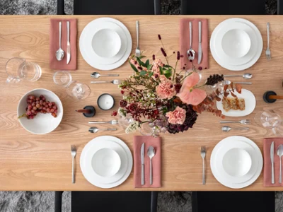 Mise en table florale et raisins Une table à manger en bois avec des assiettes blanches, des bols, des couverts sur des serviettes roses, deux bougies, un bol de raisins et une composition florale.