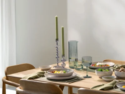 Une table en bois avec de la vaisselle Perlemor et des chandeliers Like Home Une table de salle à manger en bois avec des assiettes pleines de nourriture, des verres, des bougies vertes et de la vaisselle minimaliste.