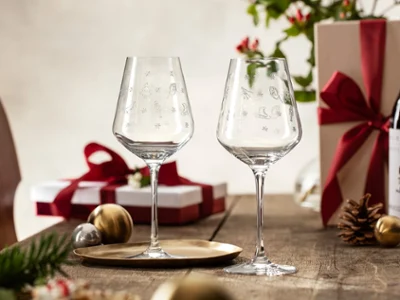 Des verres Toy’s Delight avec un emballage cadeau en arrière-plan Deux verres à vin Toy’s Delight de Villeroy & Boch sont posés sur une table au milieu de cadeaux emballés, d’une bouteille de vin et de décorations festives.