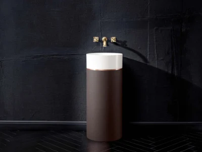 Un lavabo Octagon rond blanc sur un socle noir Un lavabo moderne avec une base cylindrique noire et une cuve blanche, ainsi qu’un robinet et des poignées en laiton, se dresse devant un mur noir structuré et un sol foncé à chevrons.