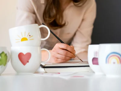 Des tasses décorées avec soin et une femme qui peint Des tasses With Love avec différents motifs, tandis qu’une femme peint en arrière-plan.