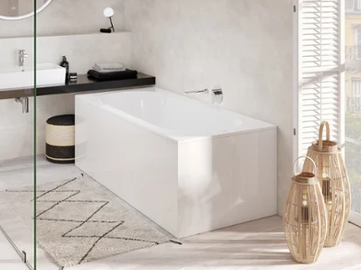 Une baignoire murale Oberon en blanc raffiné Une salle de bains moderne avec une baignoire blanche Oberon, un miroir rond au-dessus du lavabo, un tapis à motifs géométriques et une lumière naturelle entrant par une grande fenêtre. Deux lanternes en forme de panier sont placées à côté de la baignoire.