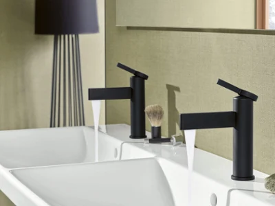Une robinetterie de lavabo Dawn noire avec un lavabo Avento Une salle de bains moderne avec deux robinets noirs au-dessus d’un lavabo blanc. Un blaireau et une serviette sont posés sur la tablette.