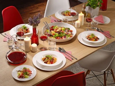 Une table de salle à manger avec des assiettes For me pleines de salade et de pâtes Une table de salle à manger avec des assiettes For me pleines de salade, de pâtes, des verres de vin rouge, des bougies et une carafe de vin rouge.