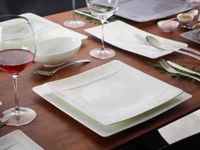 De la vaisselle Modern Grace avec des verres et du vin rouge Le set de table Modern Grace de Villeroy & Boch se compose d’assiettes carrées, de verres à vin, de plats creux et de couverts de table disposés sur une surface en bois avec des notes de fougère. Un verre à vin rouge complète parfaitement l’ambiance.