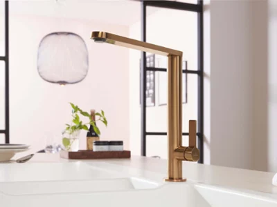 Une robinetterie de cuisine Finera en bronze sur un évier de cuisine blanc. En arrière-plan, un luminaire design est installé au-dessus d’une table. Un bocal d’herbes aromatiques est posé sur le plan de travail.