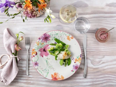 Une assiette Mariefleur et un verre à vin blanc La Divina sur une table de salle à manger avec des légumes et du poisson. Une élégante table de salle à manger avec une assiette Mariefleur à motif floral, un repas sain et un verre de vin blanc.