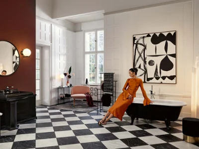 Une femme vêtue d’une robe orange est appuyée sur le bord d’une baignoire noire dans une élégante salle de bains au carrelage monochrome, avec une grande peinture abstraite et un miroir rond au-dessus d’un lavabo noir.
